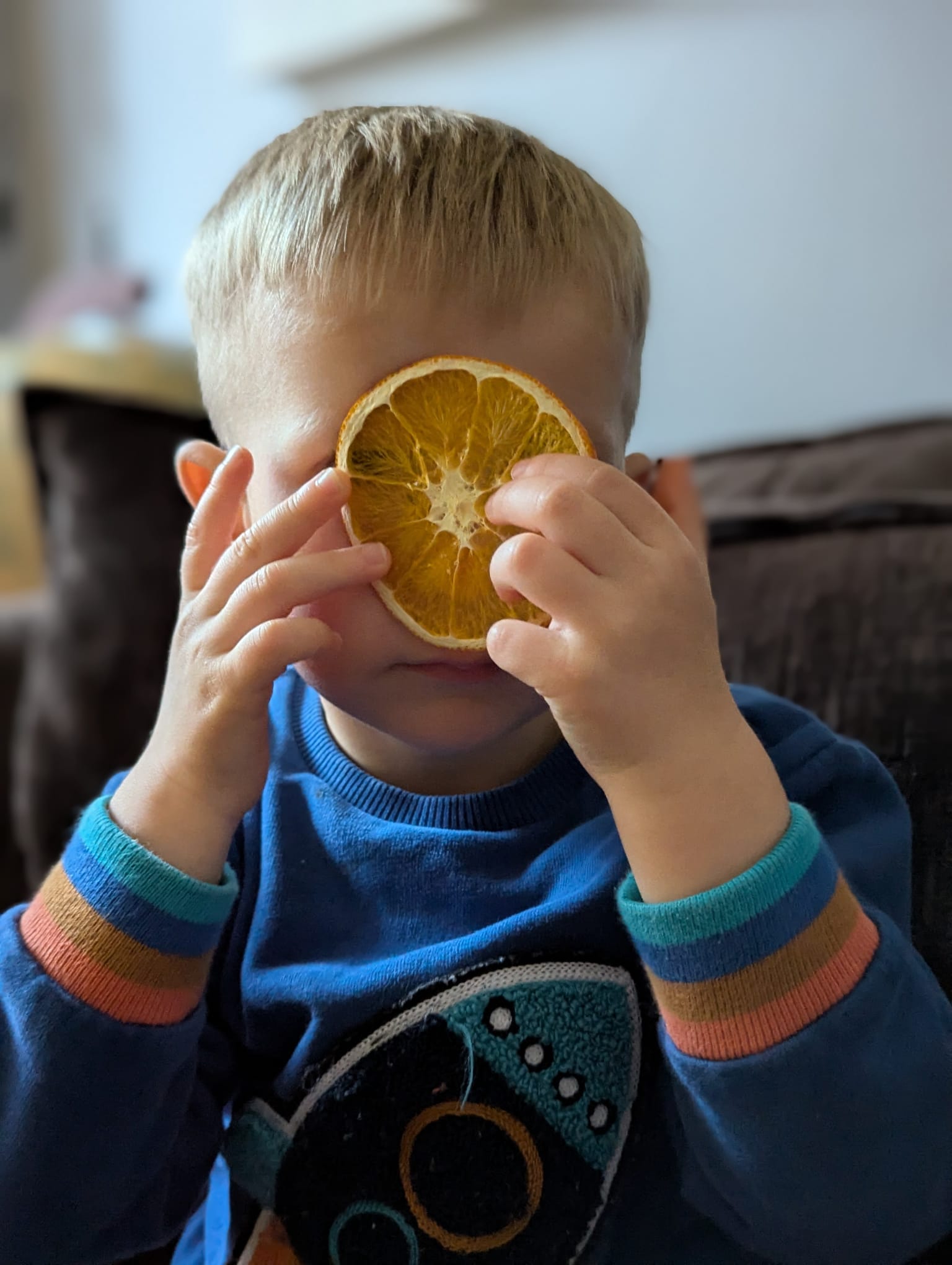 Child holding a dried orange slice over eye, playfully enjoying healthy snacks at home – fun and nutritious fruit snack for kids.