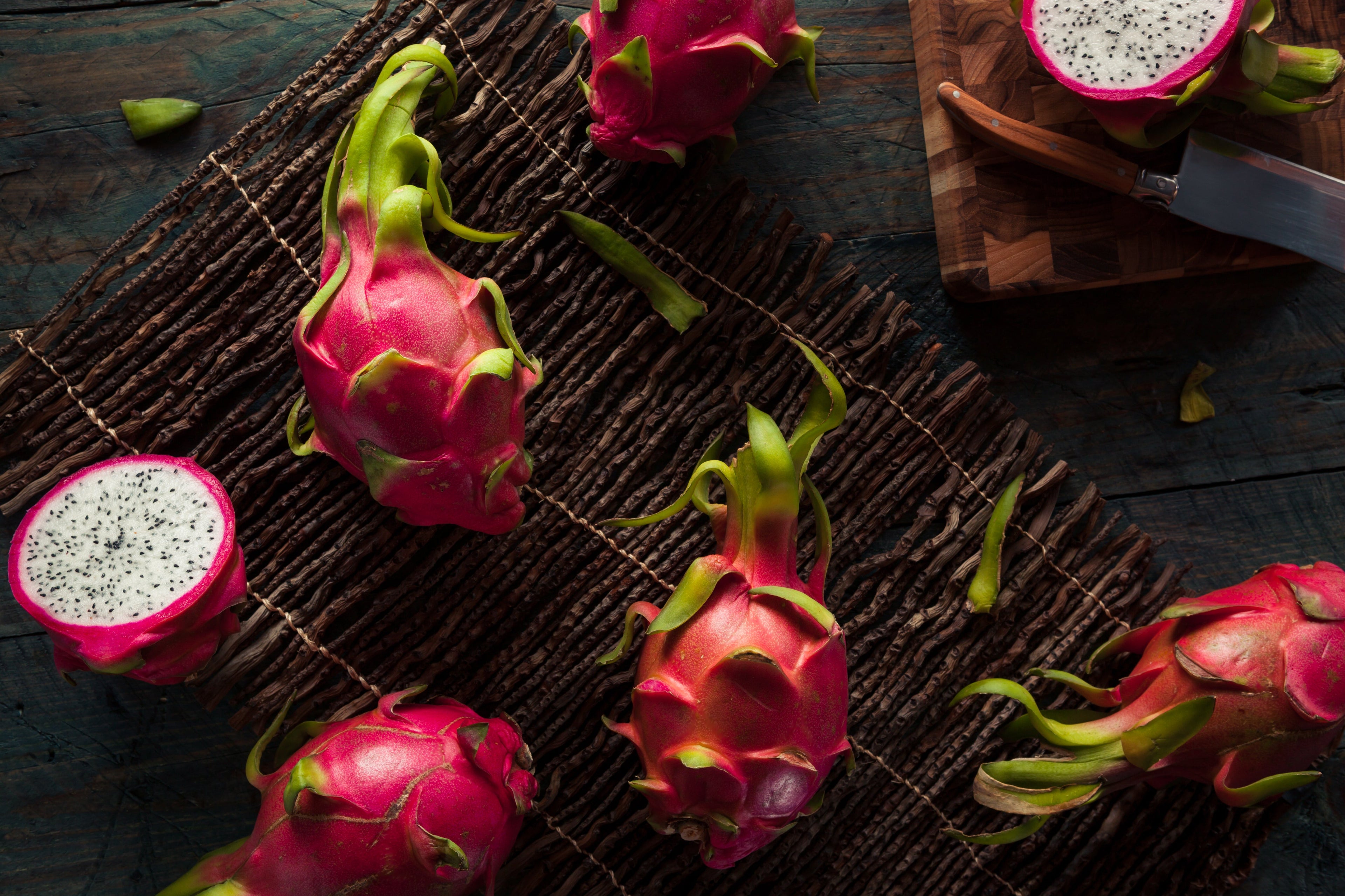 Whole and halved dragon fruits arranged on a rustic wooden surface with a knife on a chopping board, showcasing the vibrant pink skin and speckled white flesh.
