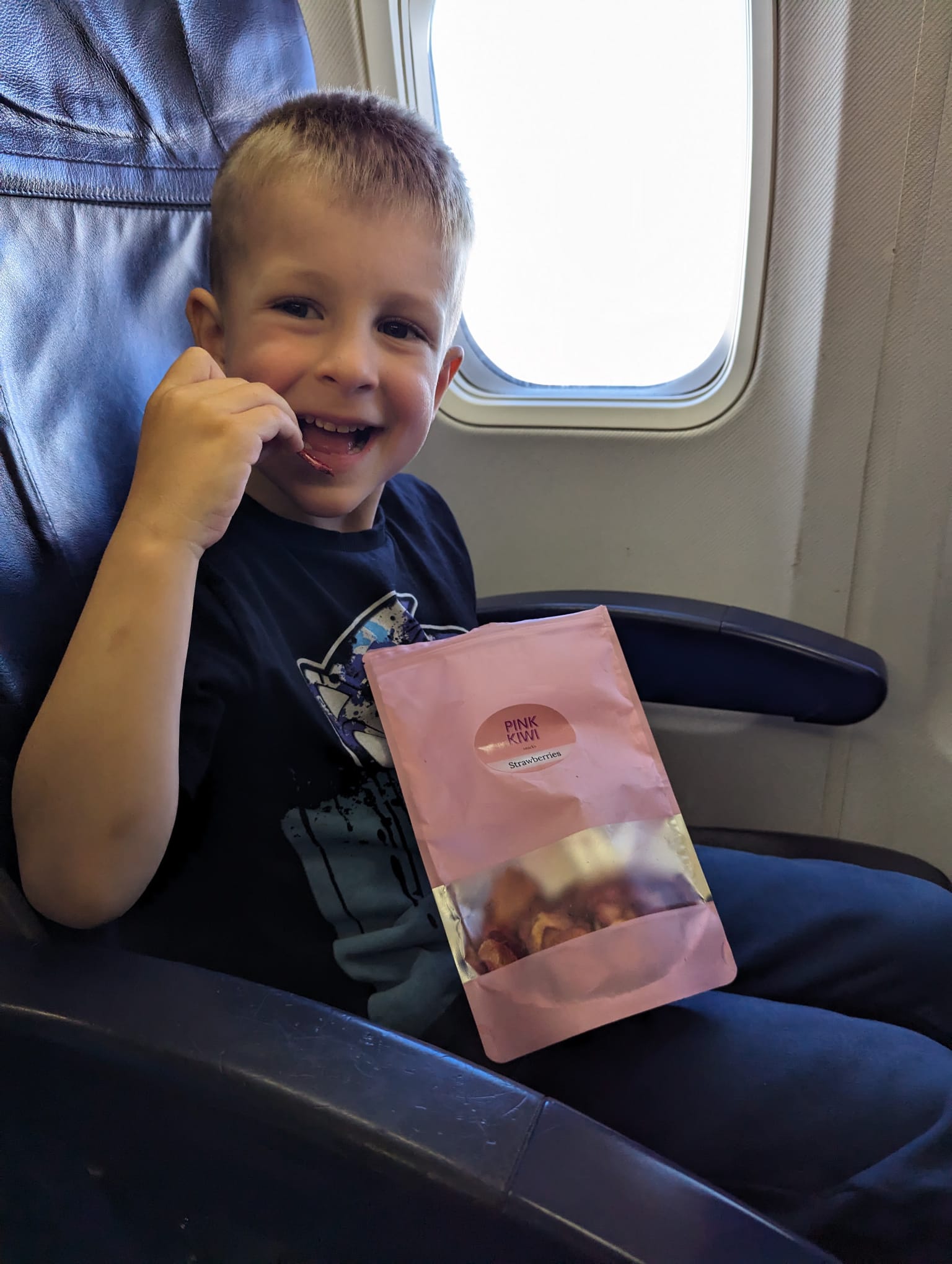 Smiling child enjoying Pink Kiwi dried strawberries while traveling on an airplane – healthy, natural snack for kids on the go.