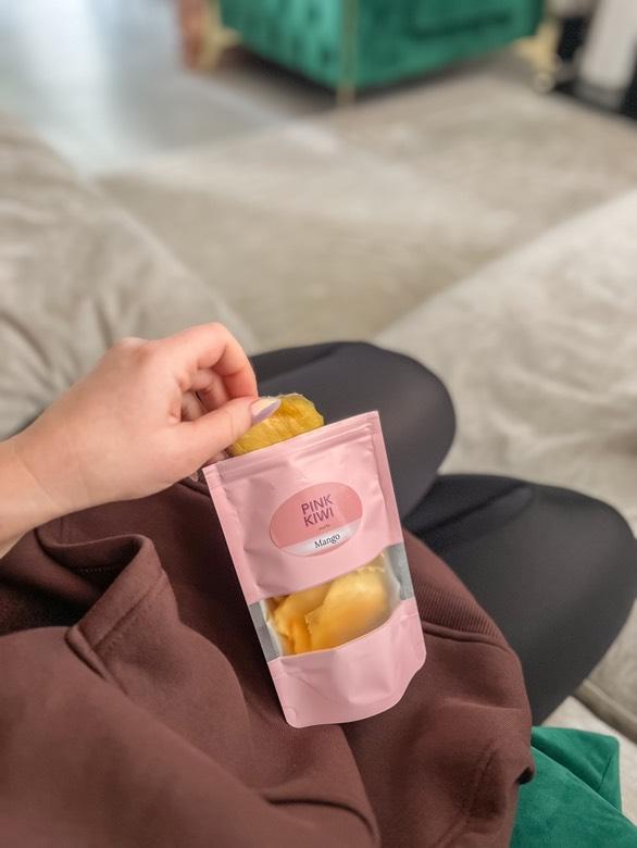Hand reaching into a Pink Kiwi Snacks pouch of dried mango while relaxing at home, highlighting a healthy and convenient snacking moment.