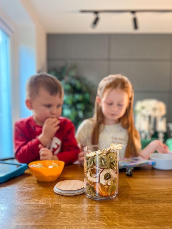 Two young children enjoying a healthy snack with a jar of assorted dried fruits on the table, promoting nutritious family-friendly snacking.