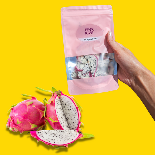 A hand holding a pink resealable pouch of Pink Kiwi Snacks dried white dragon fruit, with fresh white-fleshed dragon fruits arranged on a vibrant yellow background.