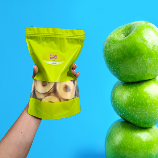Hand holding a pouch of Pink Kiwi dried apple snacks next to a stack of fresh green apples on a vibrant blue background, showcasing healthy and natural fruit snacks.