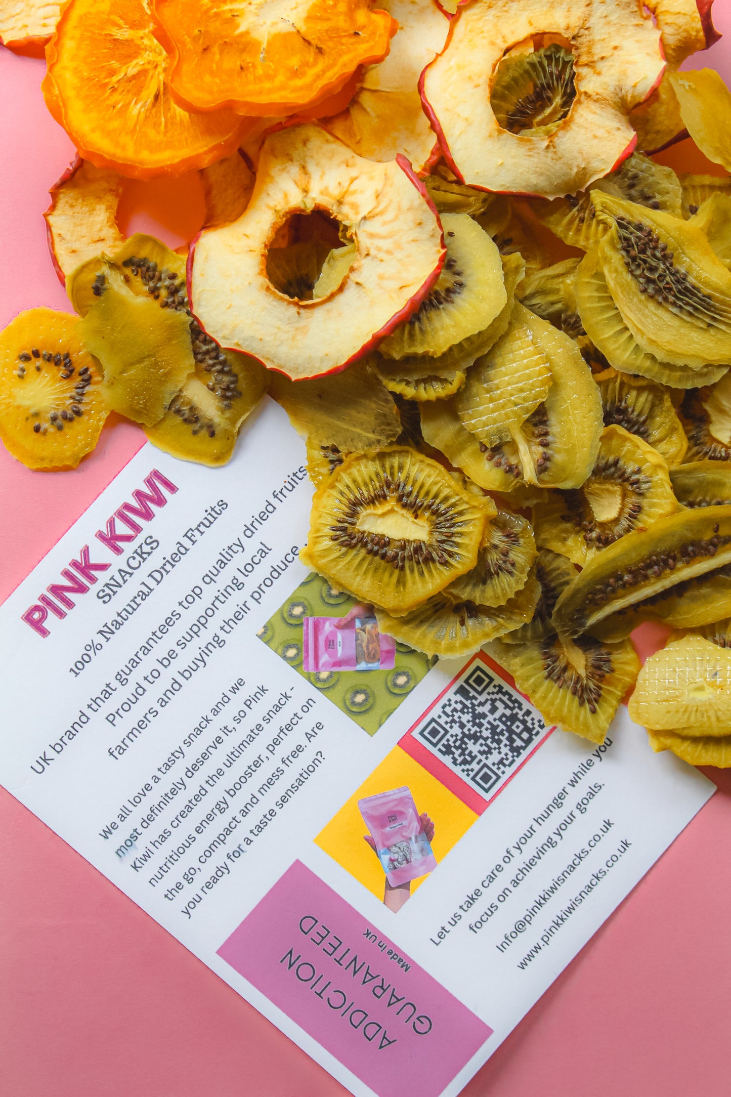 Mixed dried fruits including kiwi, apple, and mango displayed over a Pink Kiwi Snacks flyer on a pink background, highlighting brand identity and product quality.