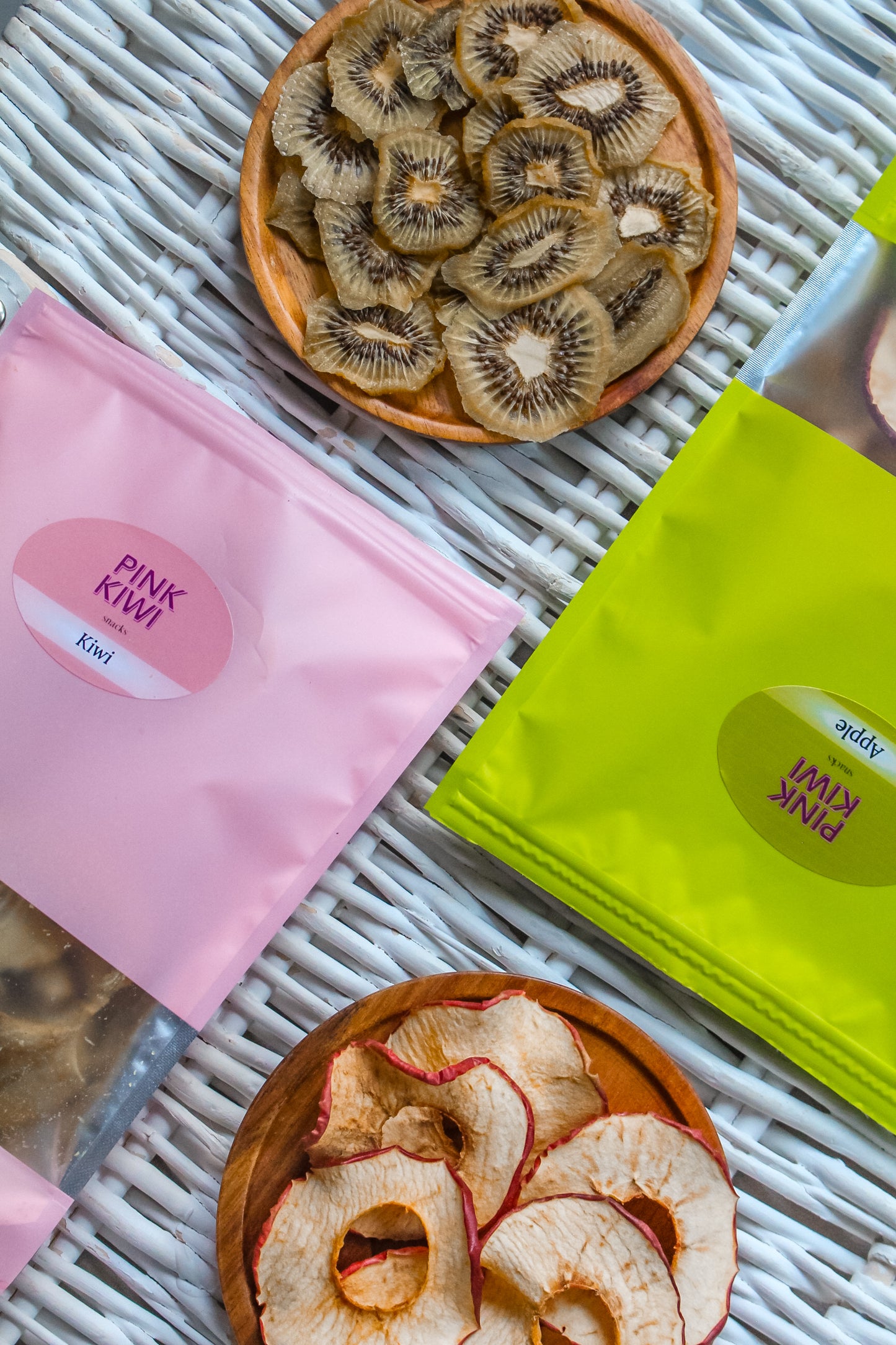 Pink Kiwi Snacks dried fruit pouches with apple and kiwi chips displayed on a white wicker surface in wooden bowls.