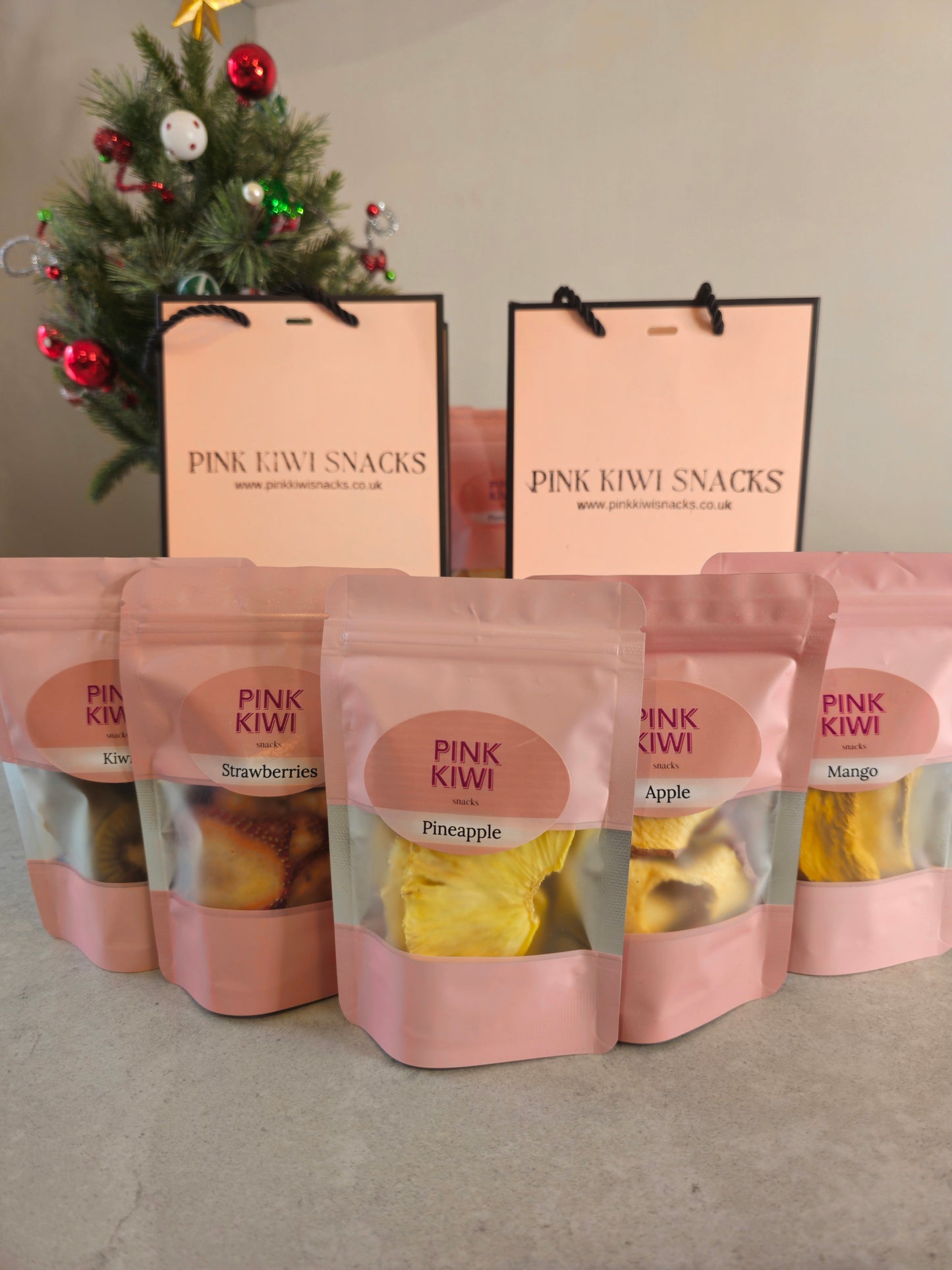 Pink Kiwi Snacks assorted dried fruit pouches including pineapple, mango, apple, and strawberries displayed in front of branded gift bags and a decorated Christmas tree.