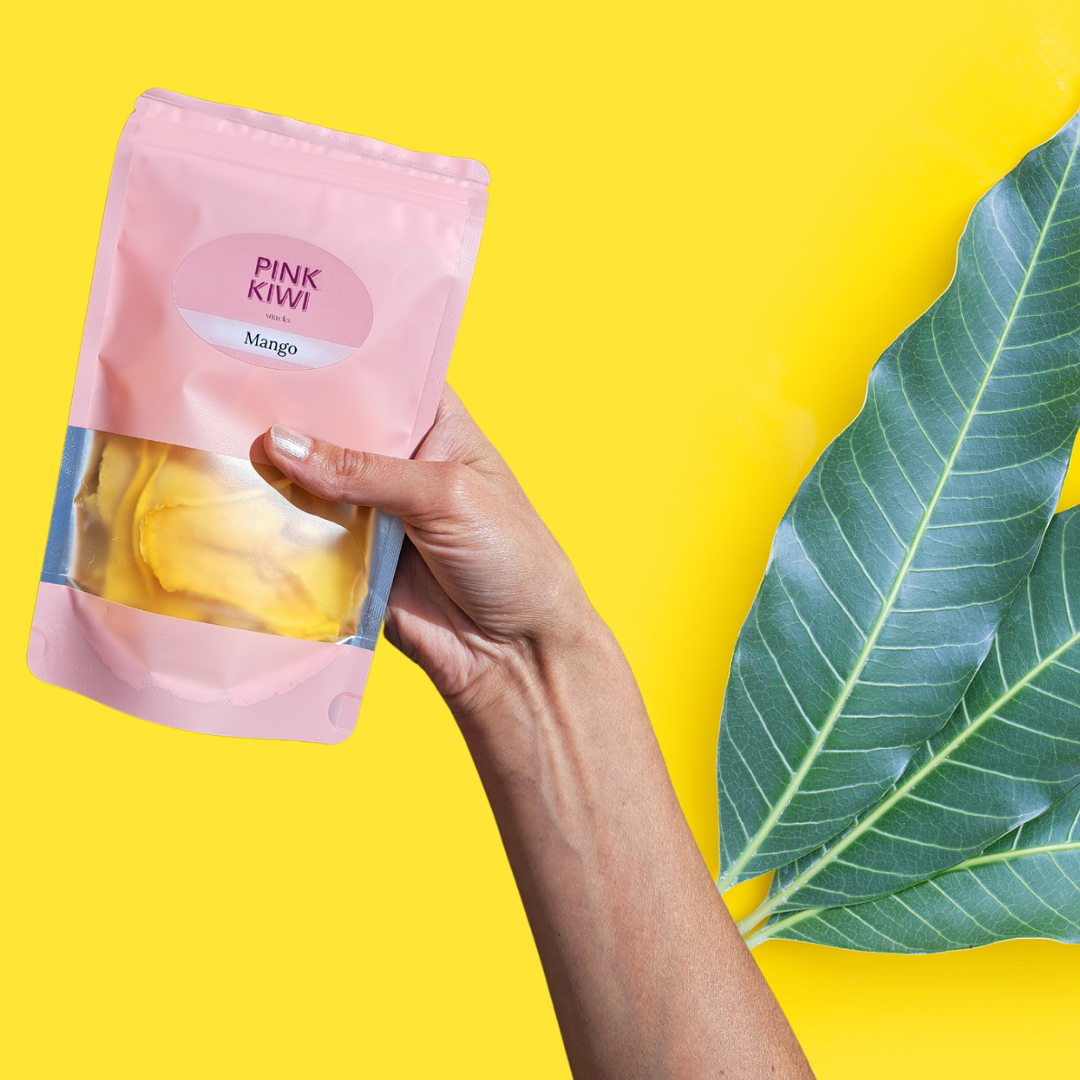 Hand holding a Pink Kiwi Snacks pouch of dried mango slices with tropical green leaves on a bright yellow background.