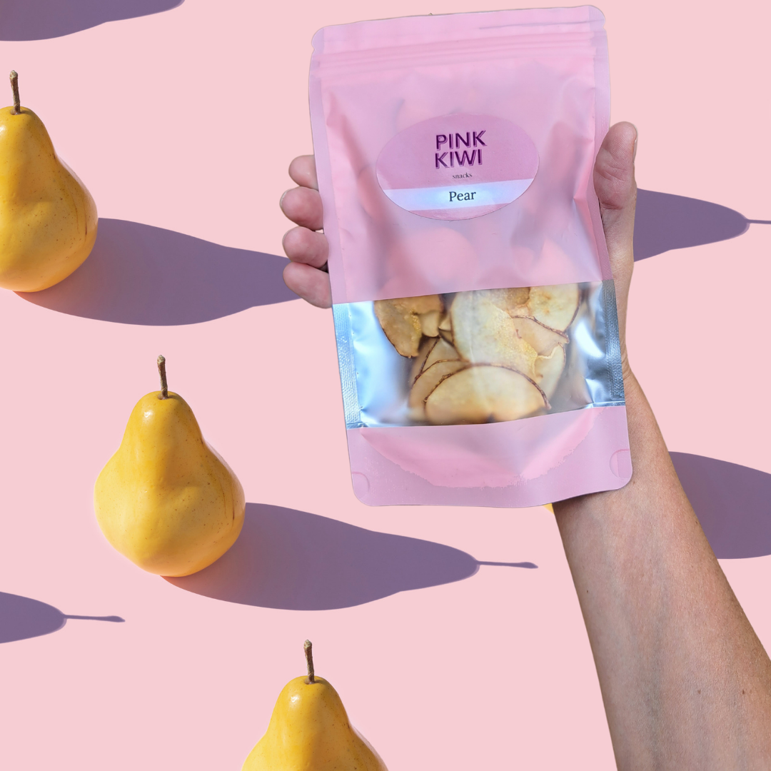 Hand holding a Pink Kiwi Snacks pouch of dried pear slices with yellow pears casting long shadows on a light pink background.