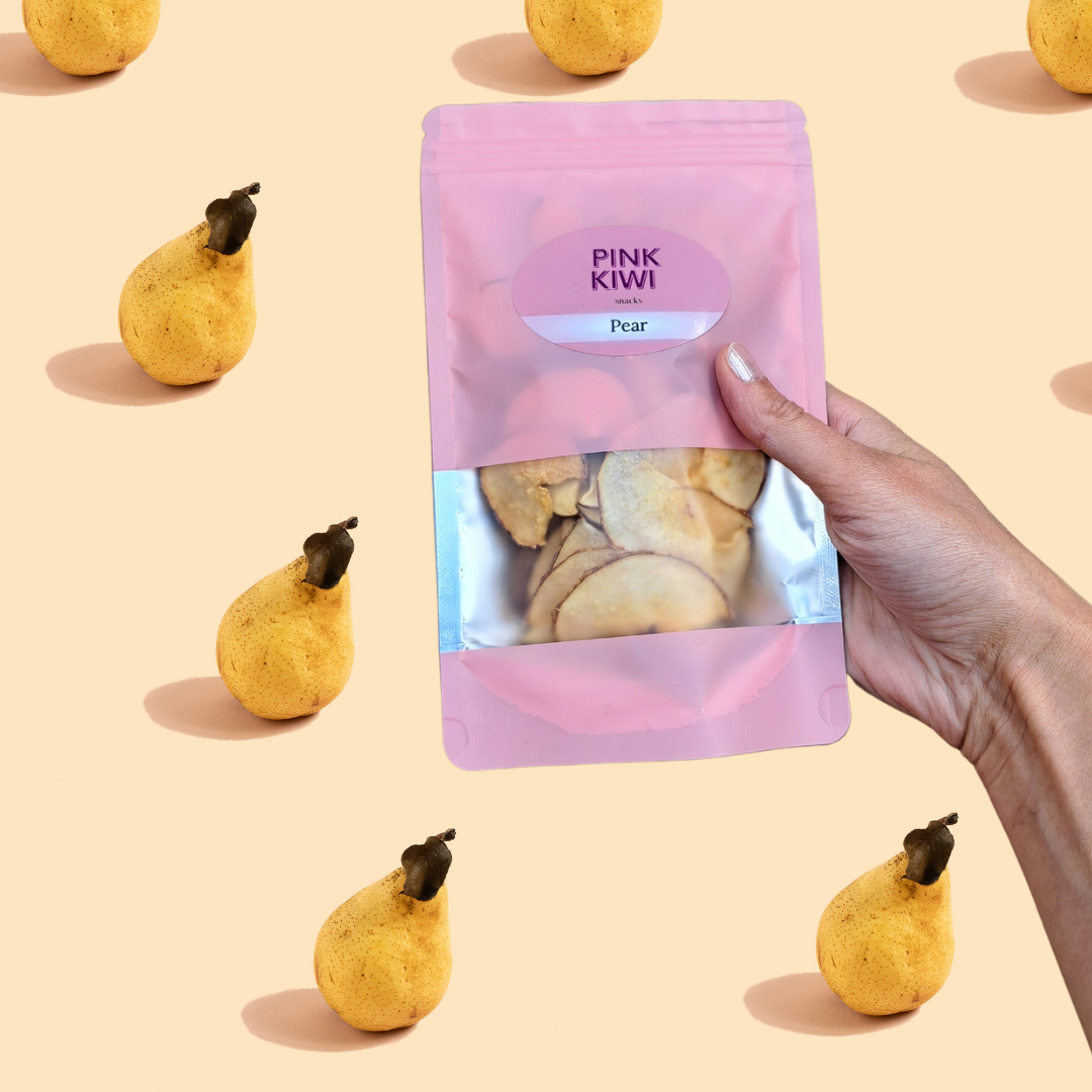 Hand holding a Pink Kiwi Snacks pouch of dried pear slices with whole pears scattered on a soft peach background.