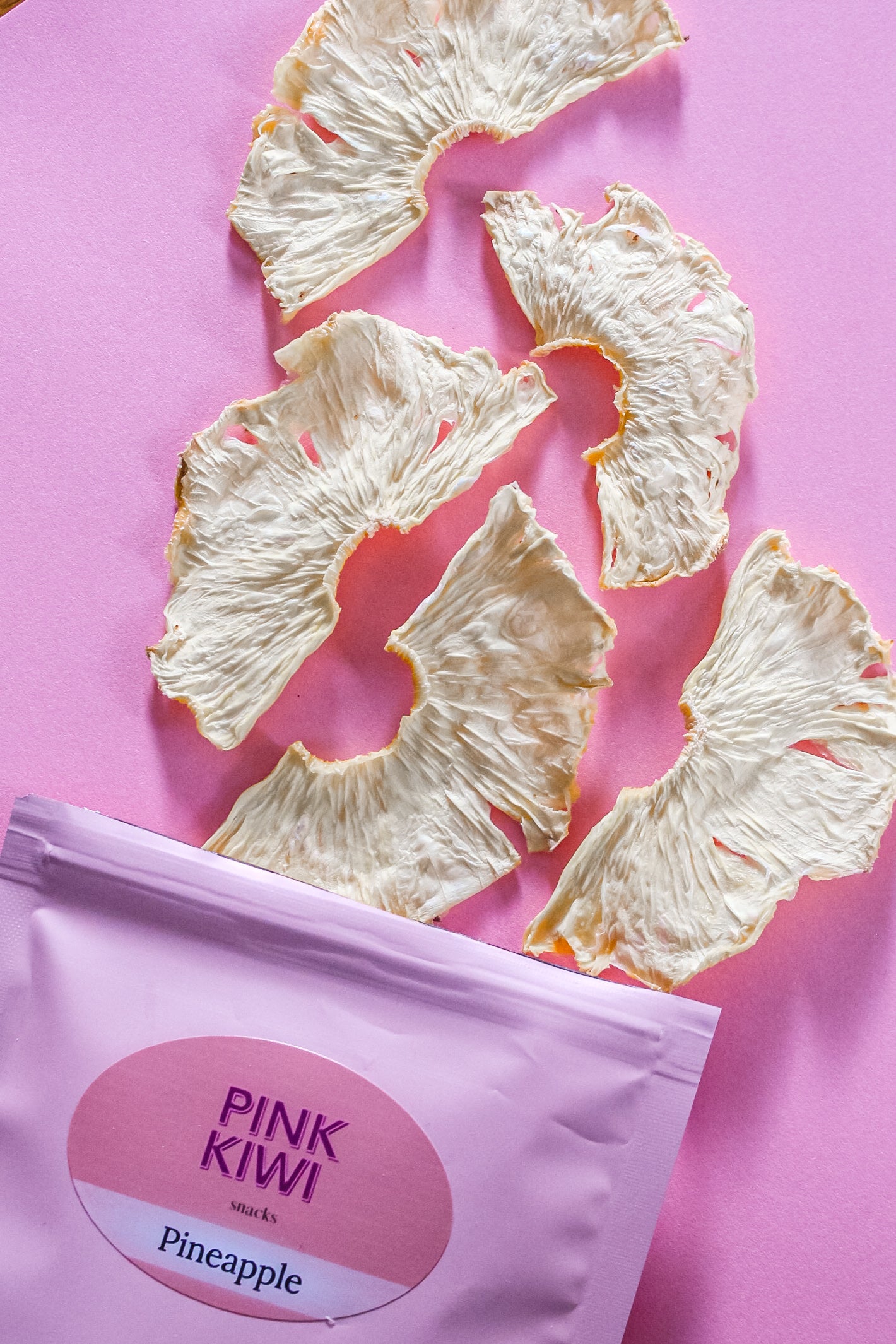 Pink Kiwi Snacks pineapple chips displayed on a pink background next to a pastel pink pouch with pineapple label.