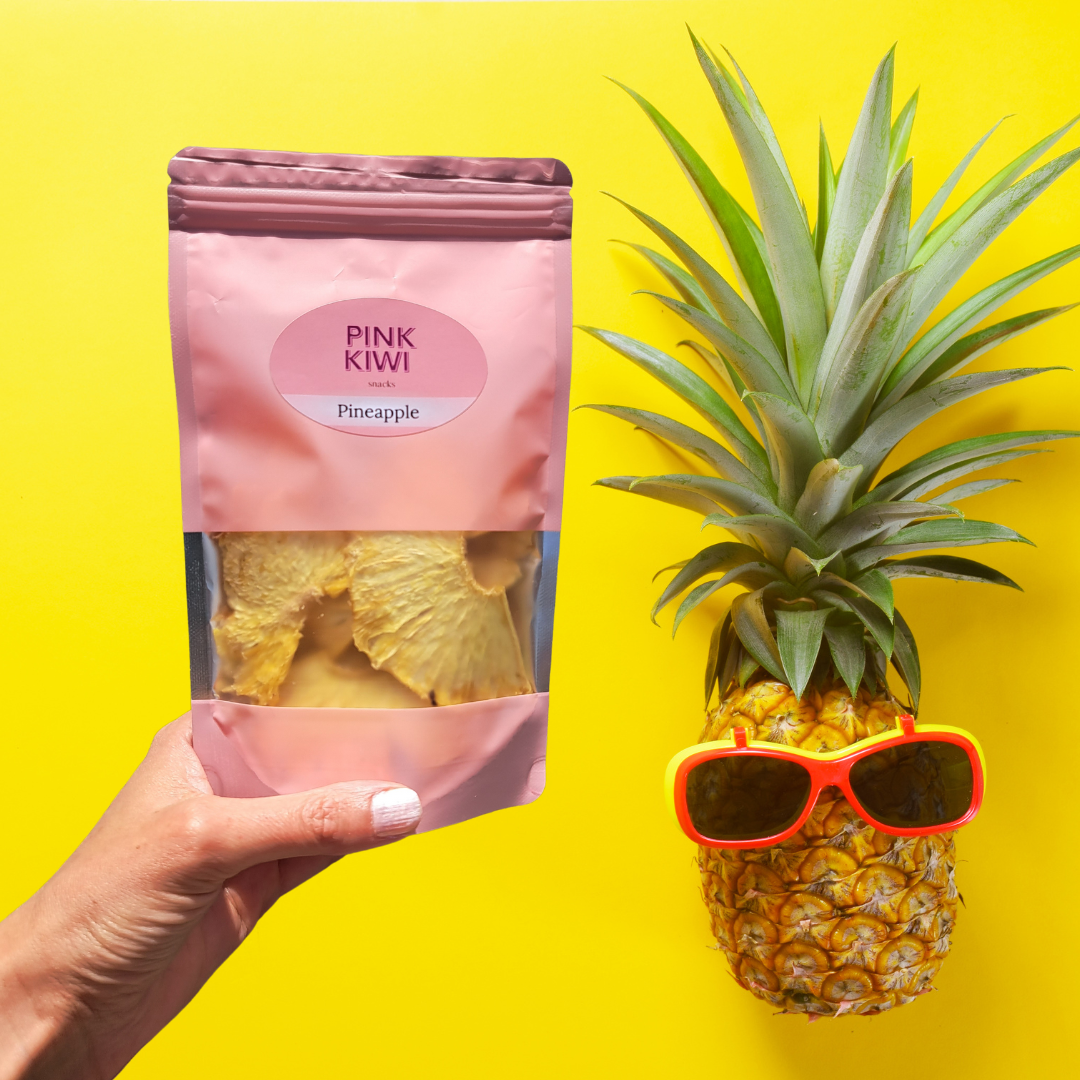Hand holding a Pink Kiwi dried pineapple snack pouch next to a fresh pineapple wearing sunglasses on a bright yellow background, highlighting tropical vibes and healthy snacking.