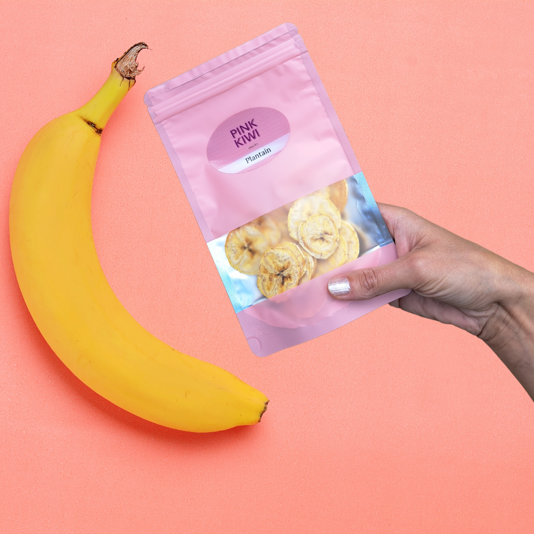 Hand holding a Pink Kiwi Snacks pouch of dried plantain slices with a fresh banana on a coral-colored background.