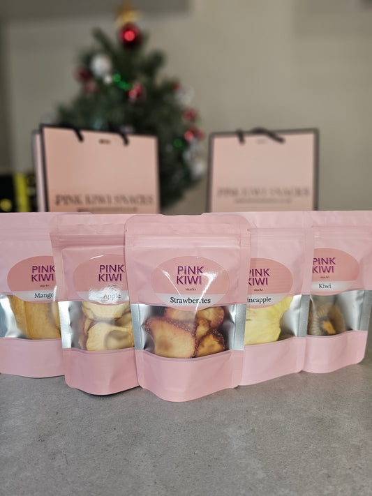 Lineup of Pink Kiwi Snacks dried fruit pouches—mango, apple, strawberries, pineapple, and kiwi—arranged in front of Christmas-themed gift bags and a decorated holiday tree.