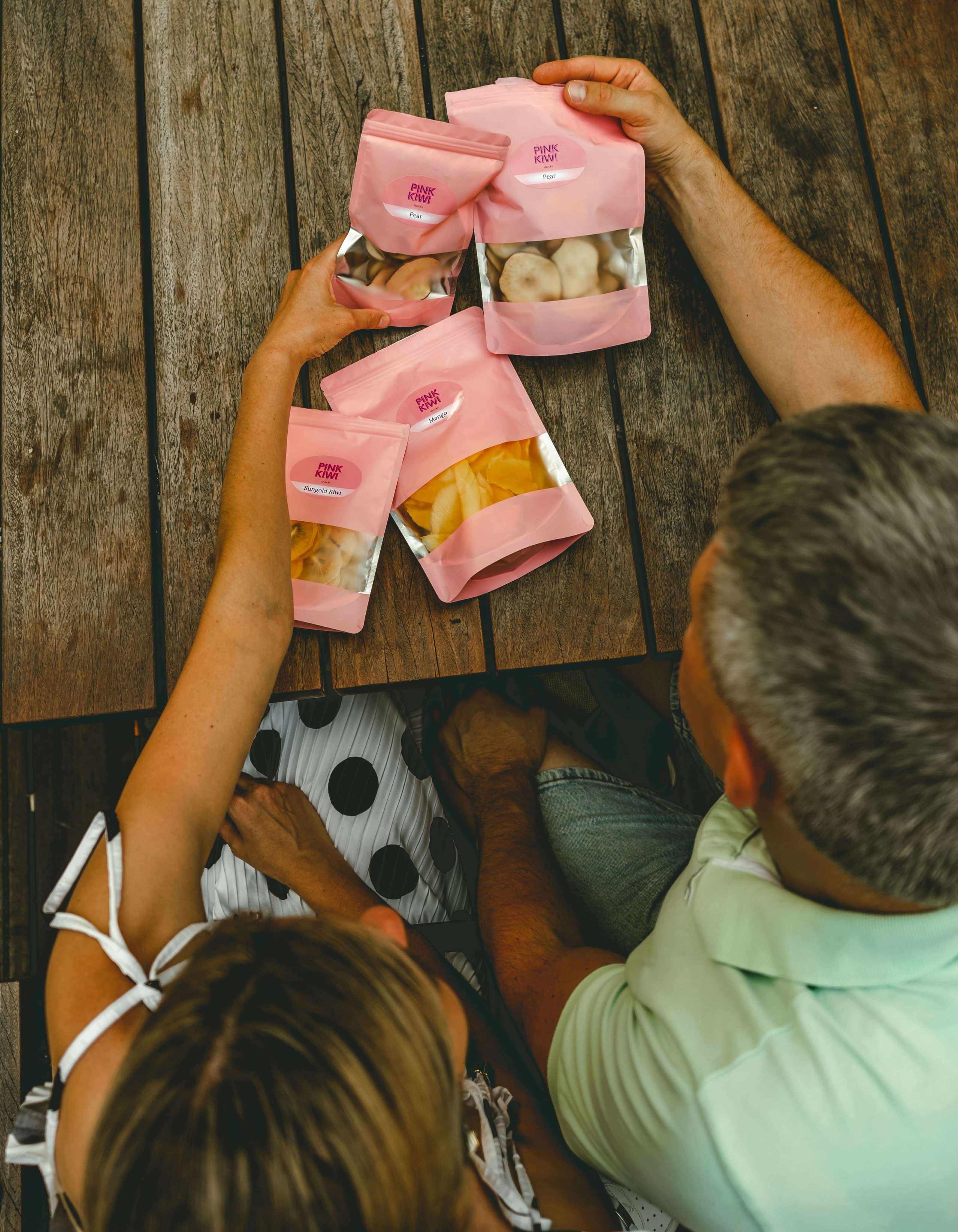 Couple enjoying and sharing Pink Kiwi Snacks' dried fruit pouches on a rustic wooden table, highlighting healthy snacking and togetherness.