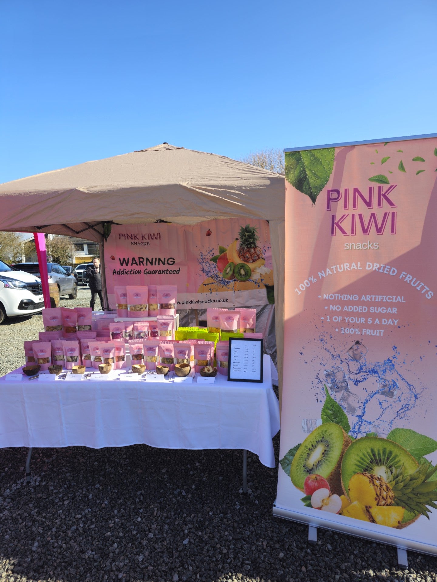 Pink Kiwi Snacks market stall at a local farmers market featuring a vibrant display of 100% natural dried fruit packs and branding.