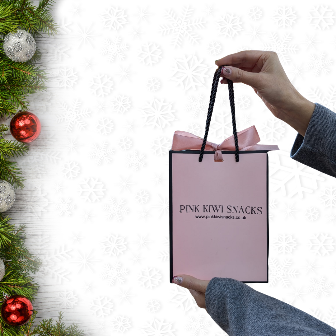 Hand holding a Pink Kiwi Snacks gift bag with a pink ribbon, set against a festive snowflake and pine background — ideal for holiday gifting.