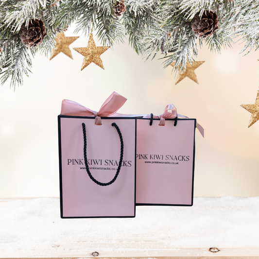 Pink Kiwi Snacks gift bags with festive ribbons, set against a snowy Christmas background with pinecones and gold stars — perfect for holiday gifting
