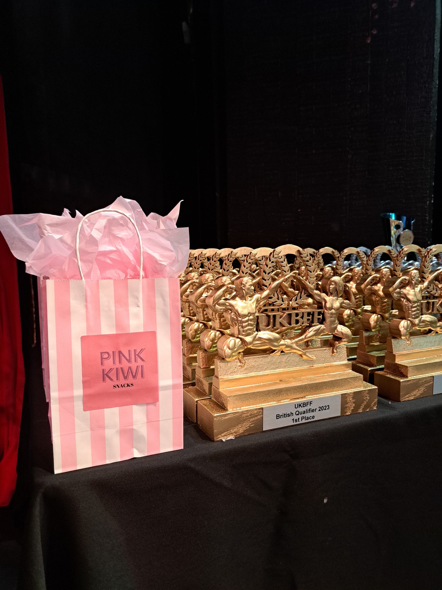 Pink Kiwi Snacks gift bag displayed beside UKBFF 2023 bodybuilding trophies at the British Qualifier event.