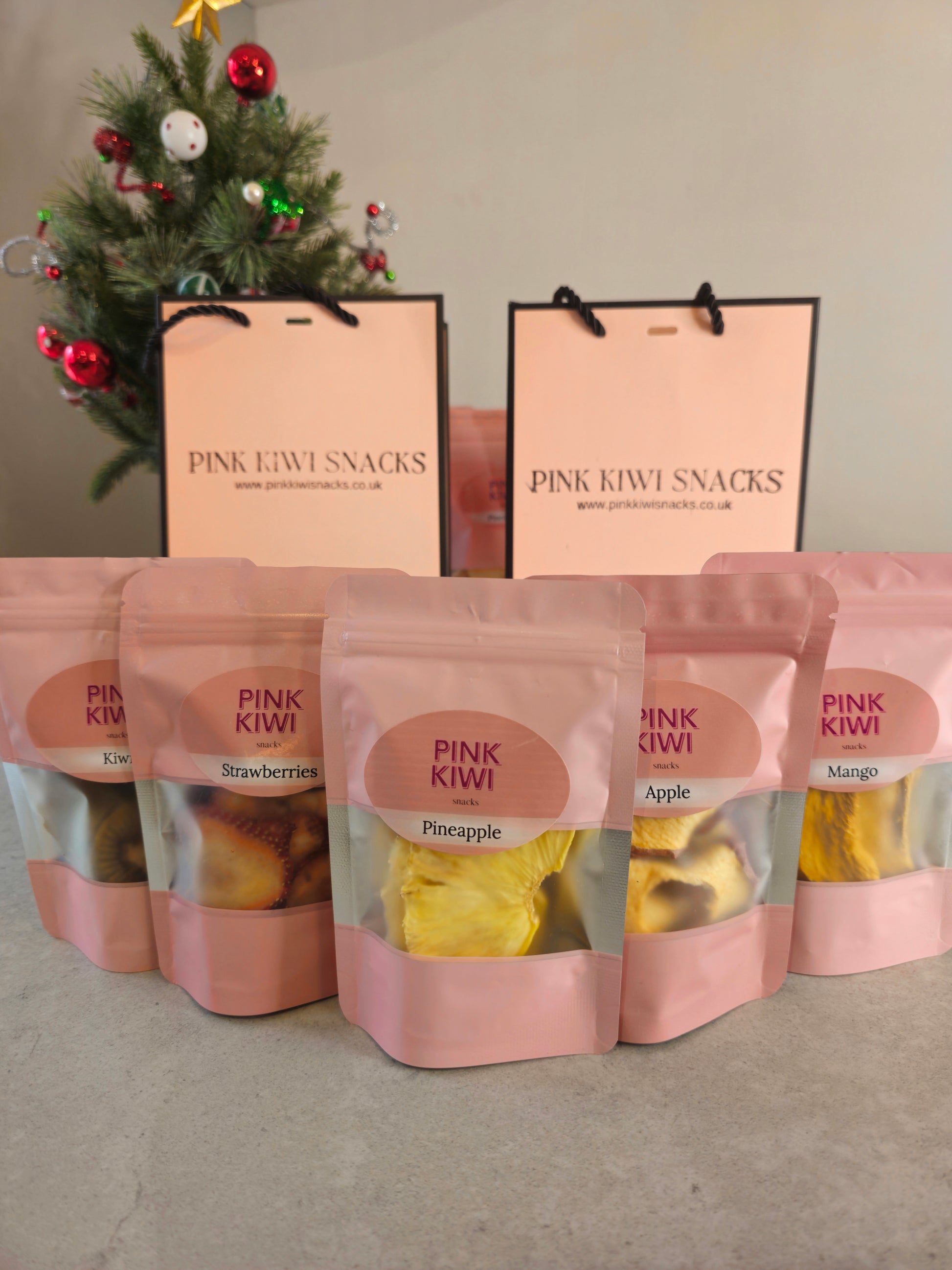 Pink Kiwi Snacks assorted dried fruit pouches including pineapple, mango, apple, and strawberries displayed in front of branded gift bags and a decorated Christmas tree.