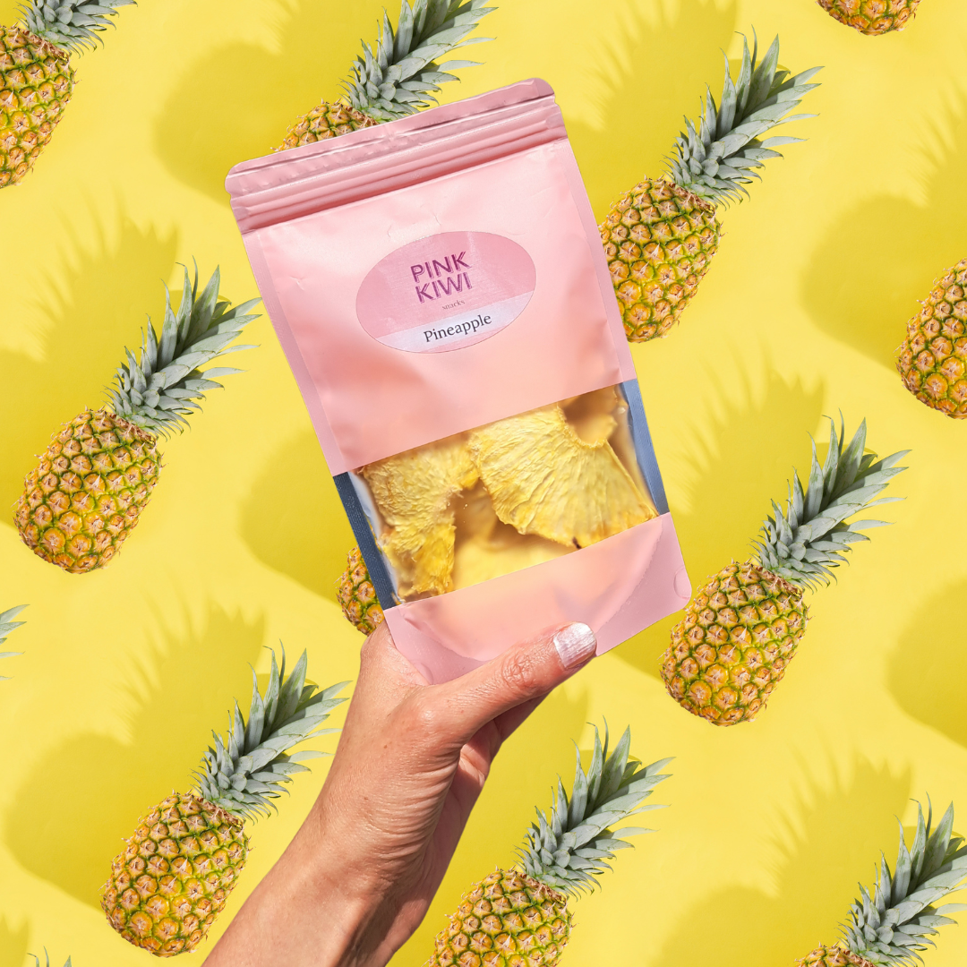 Pink Kiwi dried pineapple fruit snack held in hand against a bright yellow background with illustrated pineapples, showcasing tropical, healthy snacking vibes.