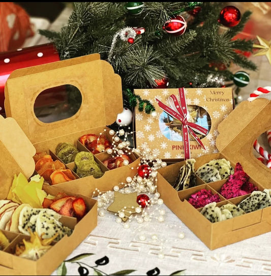 Pink Kiwi gourmet dried fruit Christmas gift boxes arranged under a festive holiday tree, featuring assorted fruit slices like dragon fruit, kiwi, persimmon, and strawberries in eco-friendly packaging.