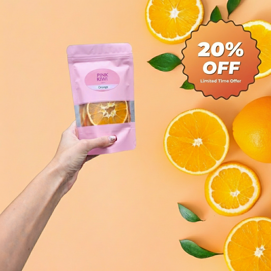 Hand holding Pink Kiwi Snacks dried orange fruit pouch with fresh orange slices and green leaves on a peach-colored background.