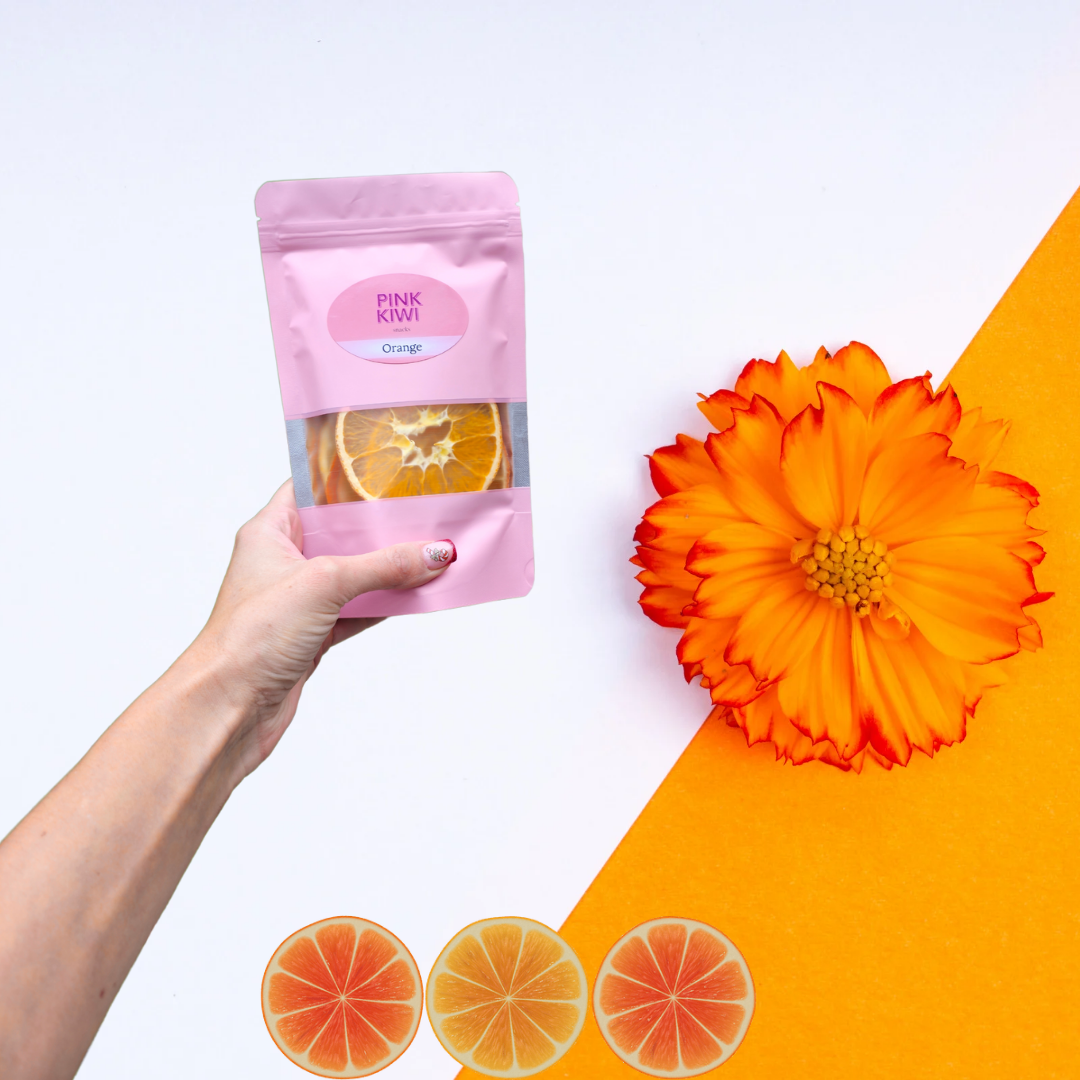 Hand holding Pink Kiwi Snacks orange dried fruit pouch on a white and orange background, decorated with an orange flower and citrus slice graphics.