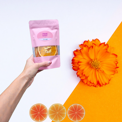 Hand holding Pink Kiwi Snacks orange dried fruit pouch on a white and orange background, decorated with an orange flower and citrus slice graphics.