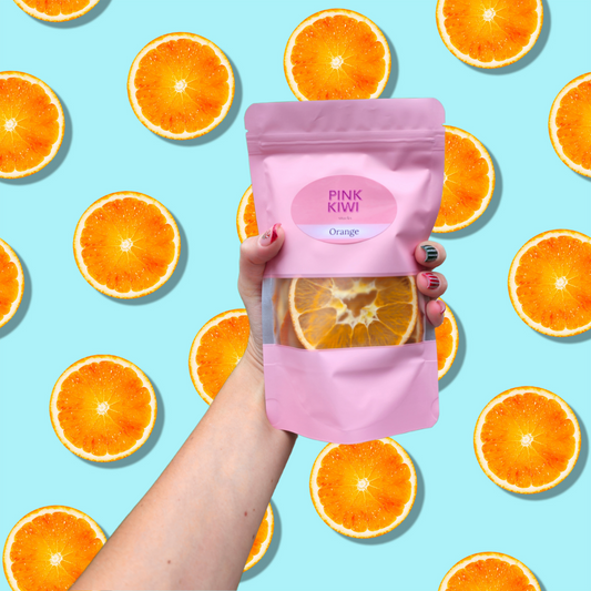 Hand holding a Pink Kiwi Snacks orange dried fruit pouch against a bright blue background with orange slices — vibrant and fresh citrus-themed design.