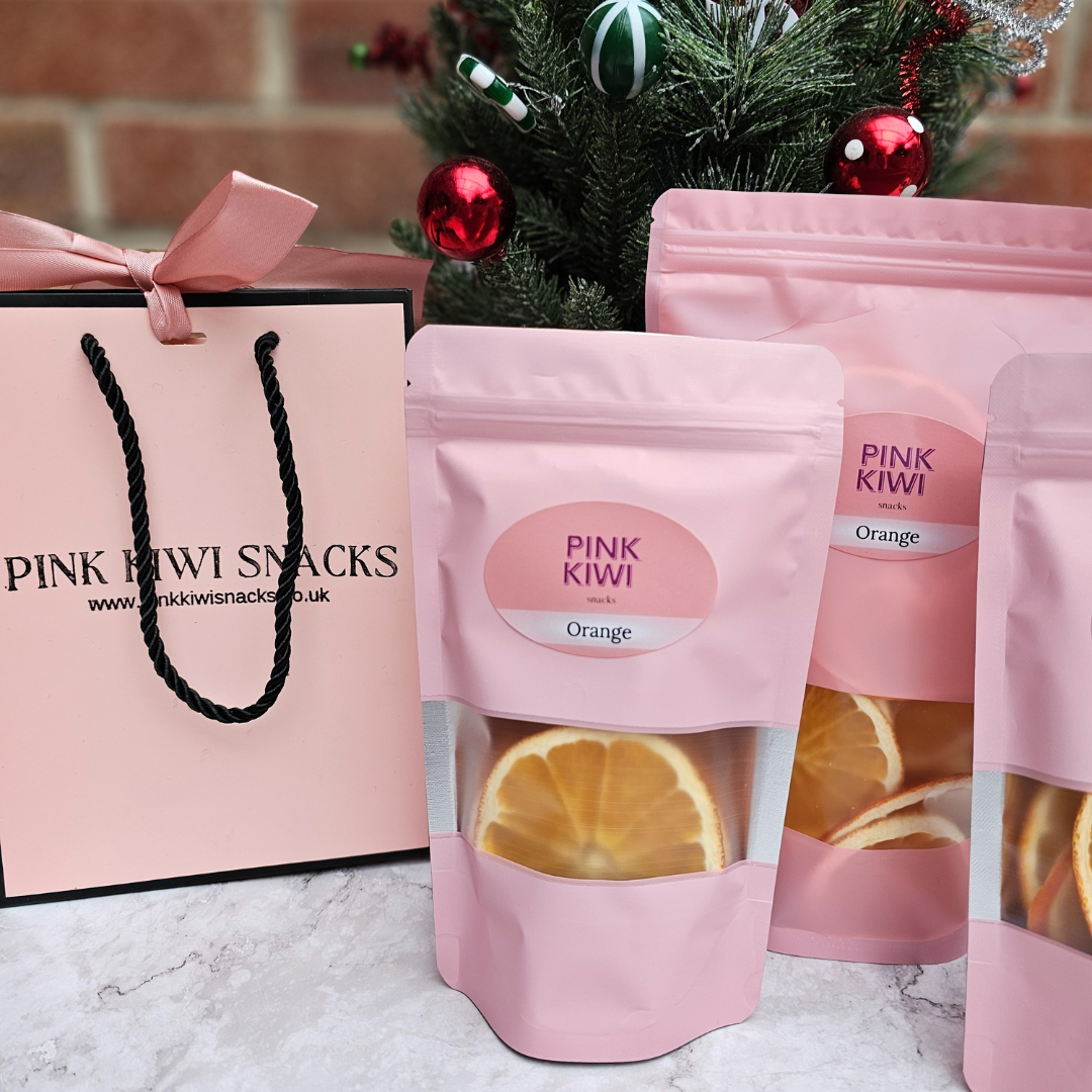 Pink Kiwi Snacks orange dried fruit packs displayed with festive Christmas tree and branded gift bag, ideal for healthy holiday gifting