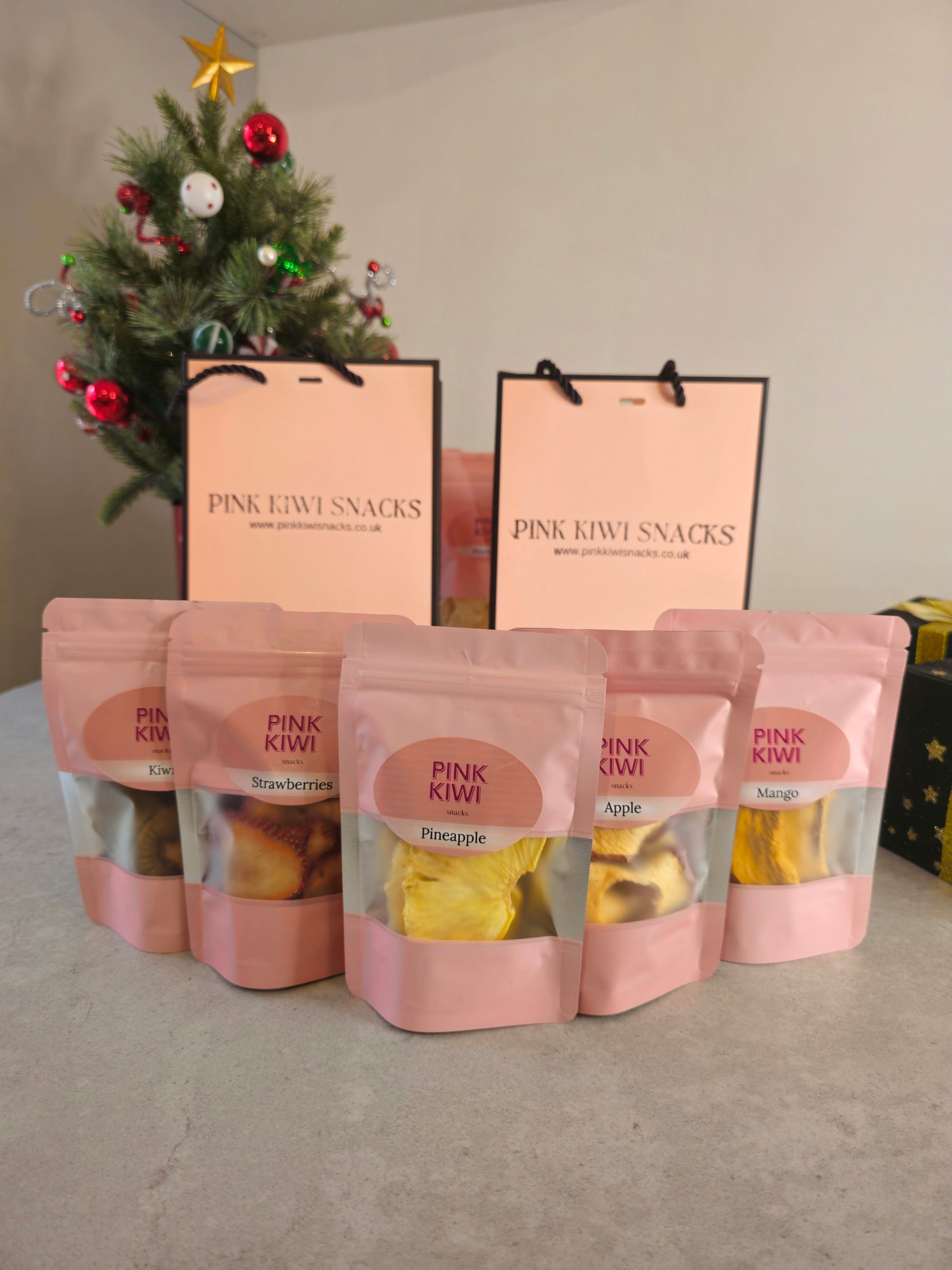 Christmas-themed display of Pink Kiwi Snacks dried fruit pouches, including kiwi, strawberries, pineapple, apple, and mango, with branded gift bags and a decorated tree in the background.