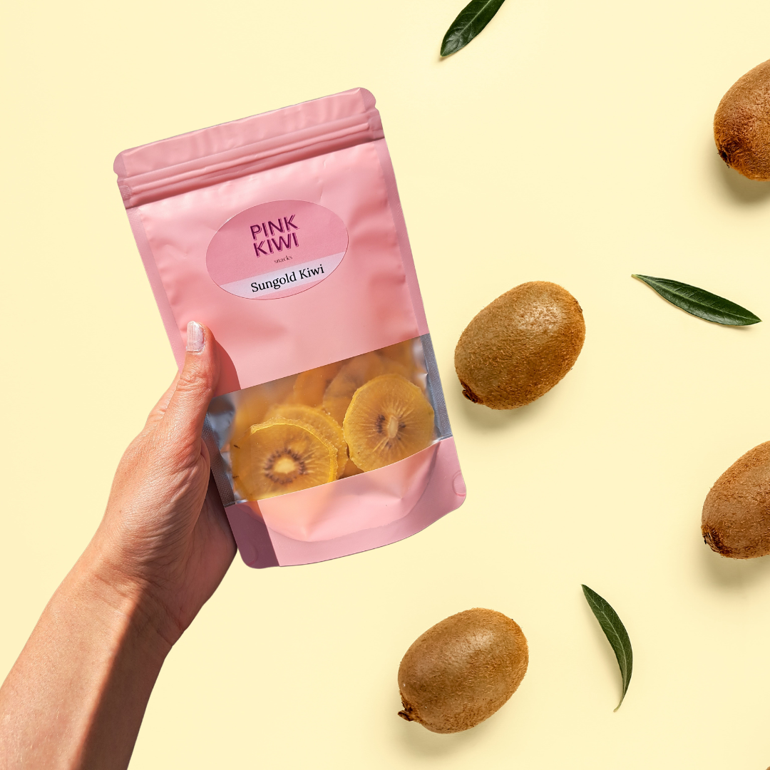 Pink Kiwi Sungold dried kiwi fruit snack pack held in hand, surrounded by whole kiwis and green leaves on a soft cream background, representing healthy tropical snacking.